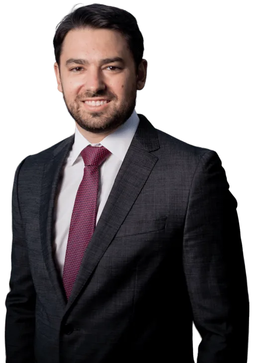 Man in a dark suit, white dress shirt, and red tie, standing and smiling slightly, facing the camera against a plain background.