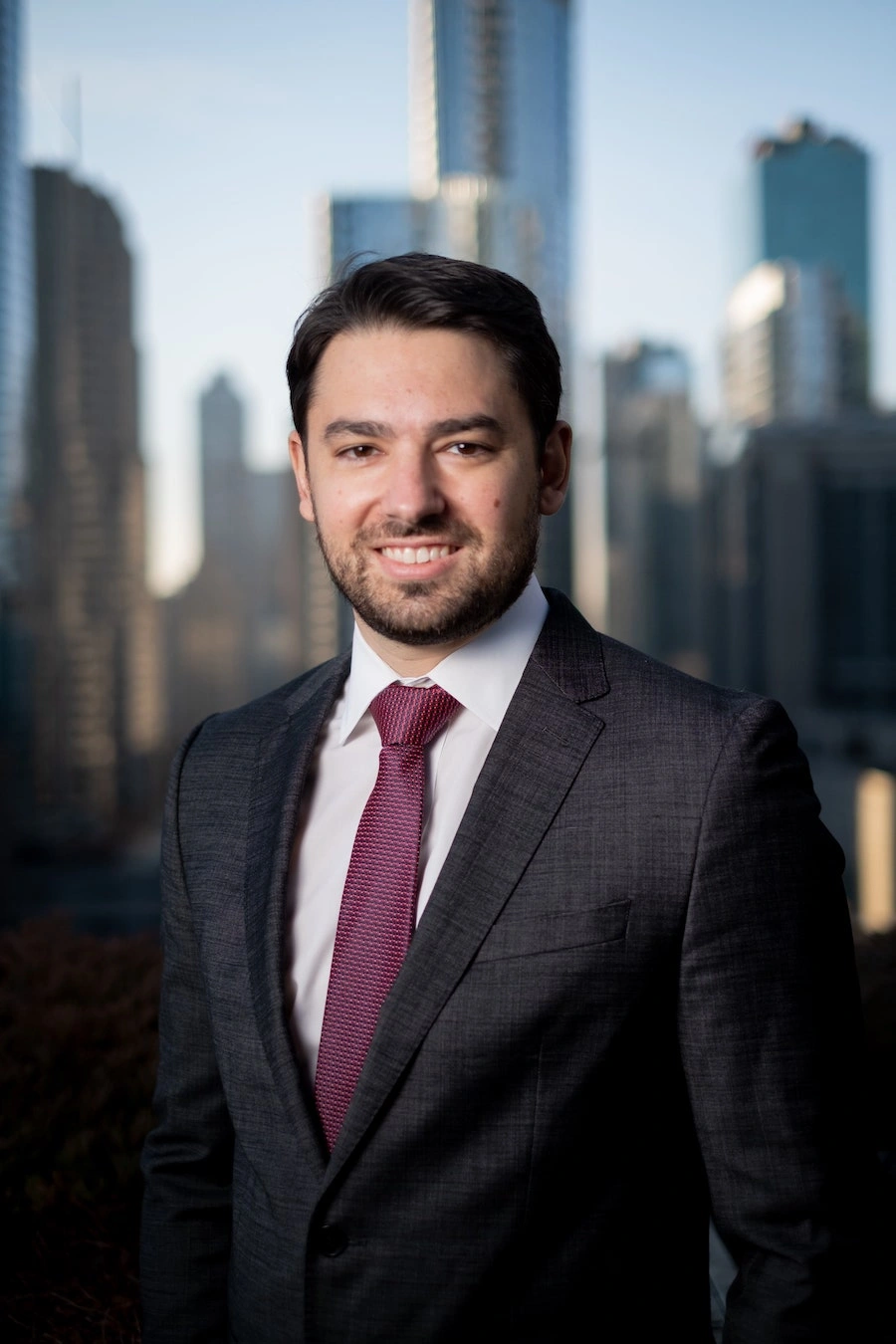 A man in a dark suit and red tie stands outdoors with a city skyline in the background.