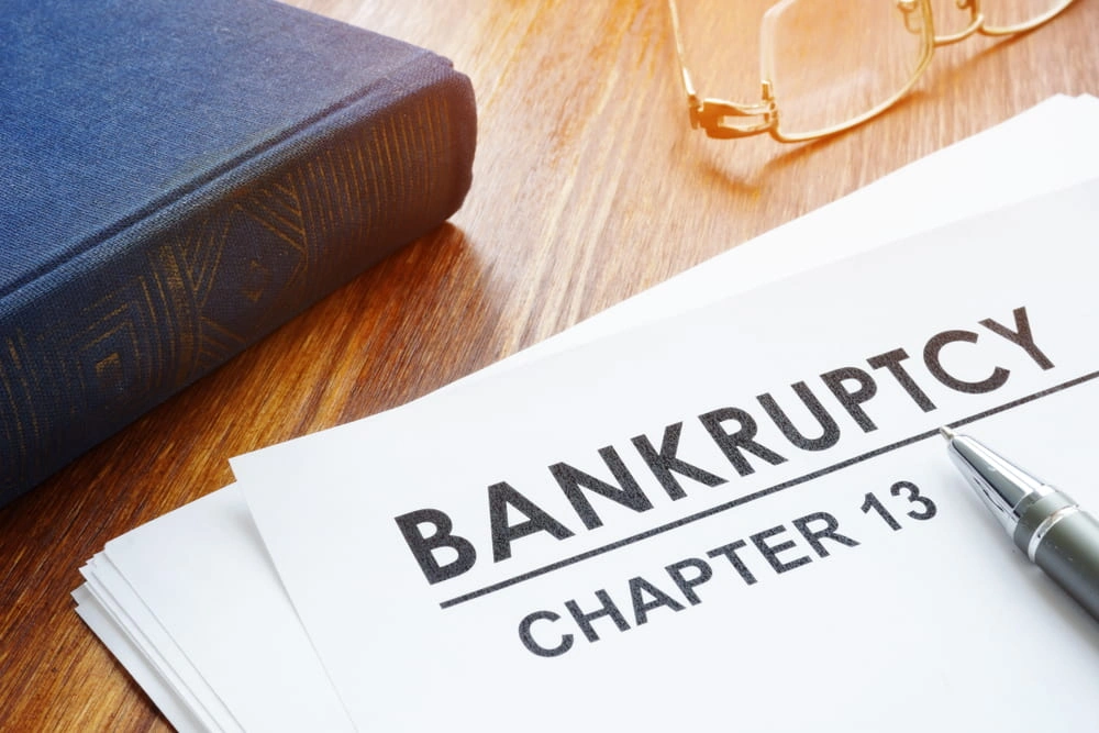 A document labeled "Bankruptcy Chapter 13" on a desk with a pen, a closed book, and a pair of eyeglasses nearby.