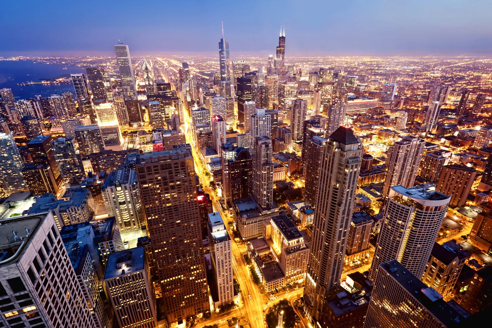 Aerial view of a city skyline at dusk with illuminated skyscrapers, busy streets, and a distant waterfront.