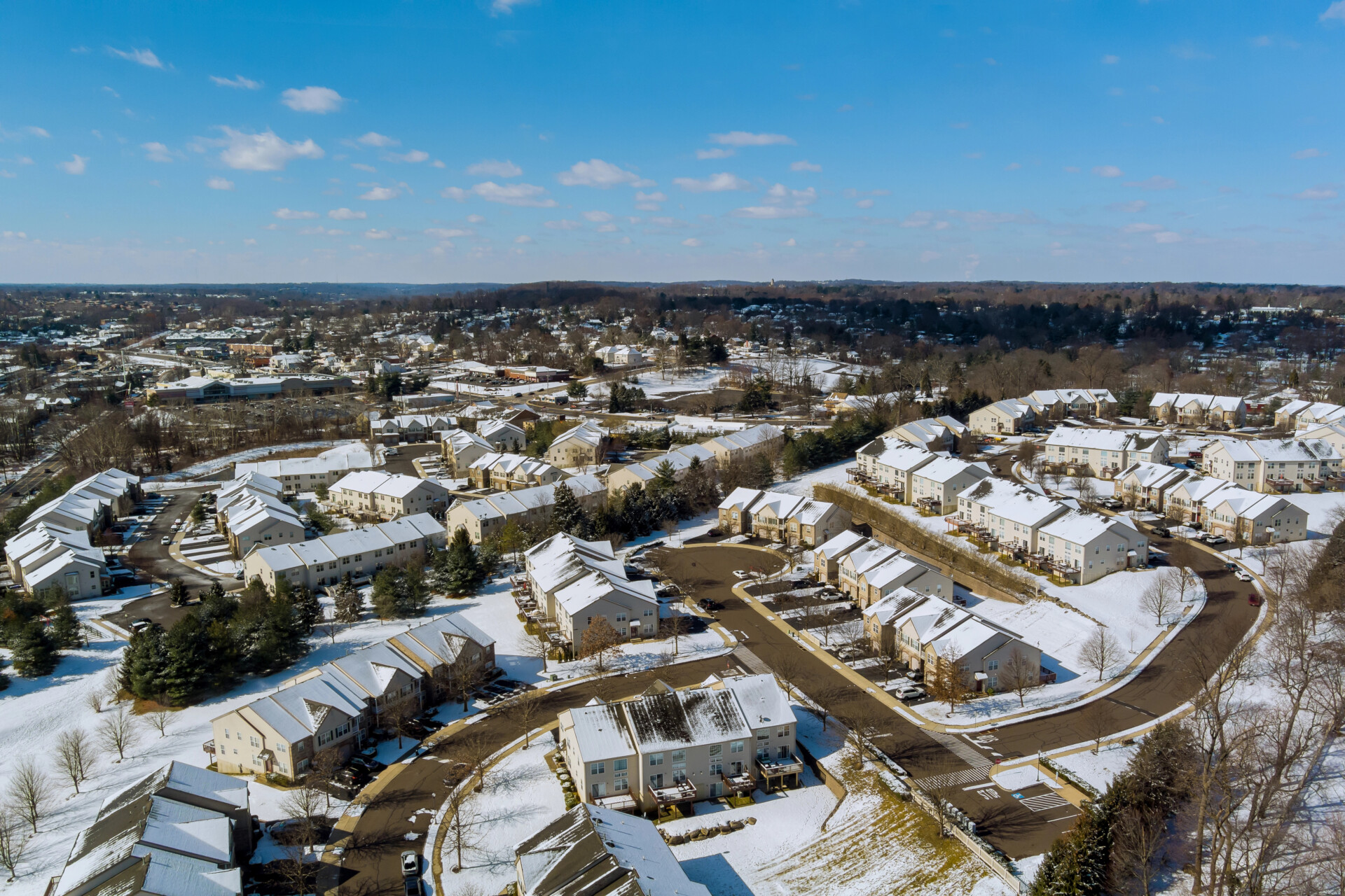 Aerial view of a suburban neighborhood with snow-covered rooftops and streets under a clear blue sky.
