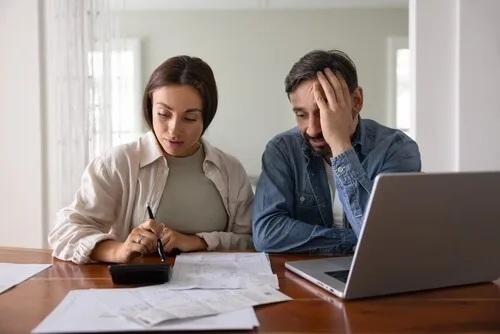 Couple reviewing documents at table with calculator and laptop, concerned expressions visible